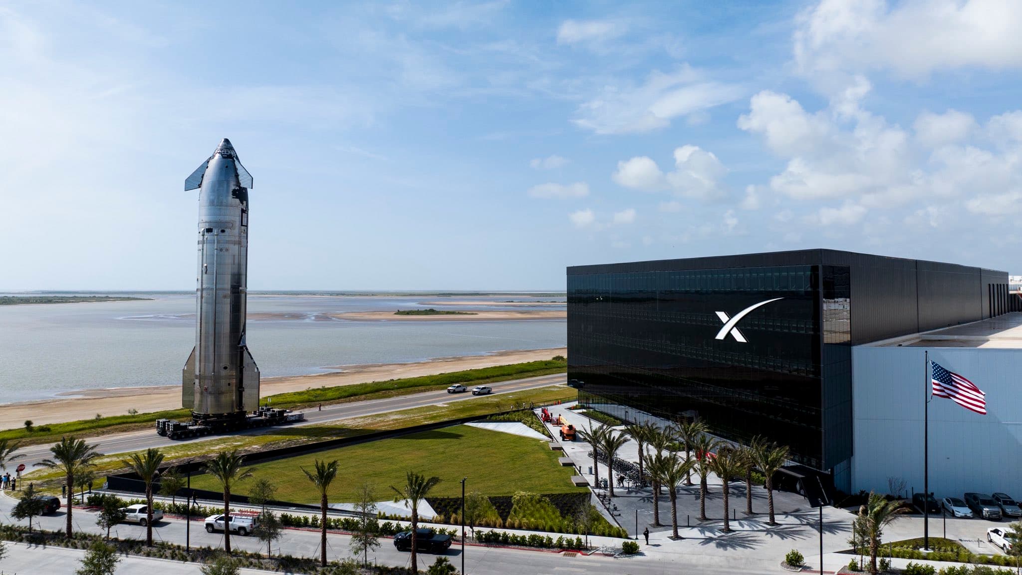 A Starship rolls past the SpaceX Starfactory at Starbase, Texas, with Boca Chica beach and the Gulf beyond.